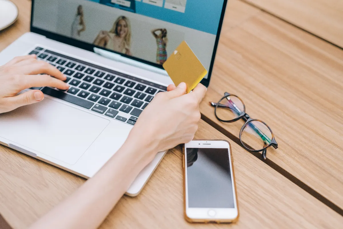 Close-up of a shopper typing on a laptop while holding a yellow payment card, with an online clothing store on screen and a phone and glasses on the desk